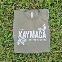 A green t-shirt with the Xaymaca Coffee Traders logo printed in the center, laid out on a grassy surface.
