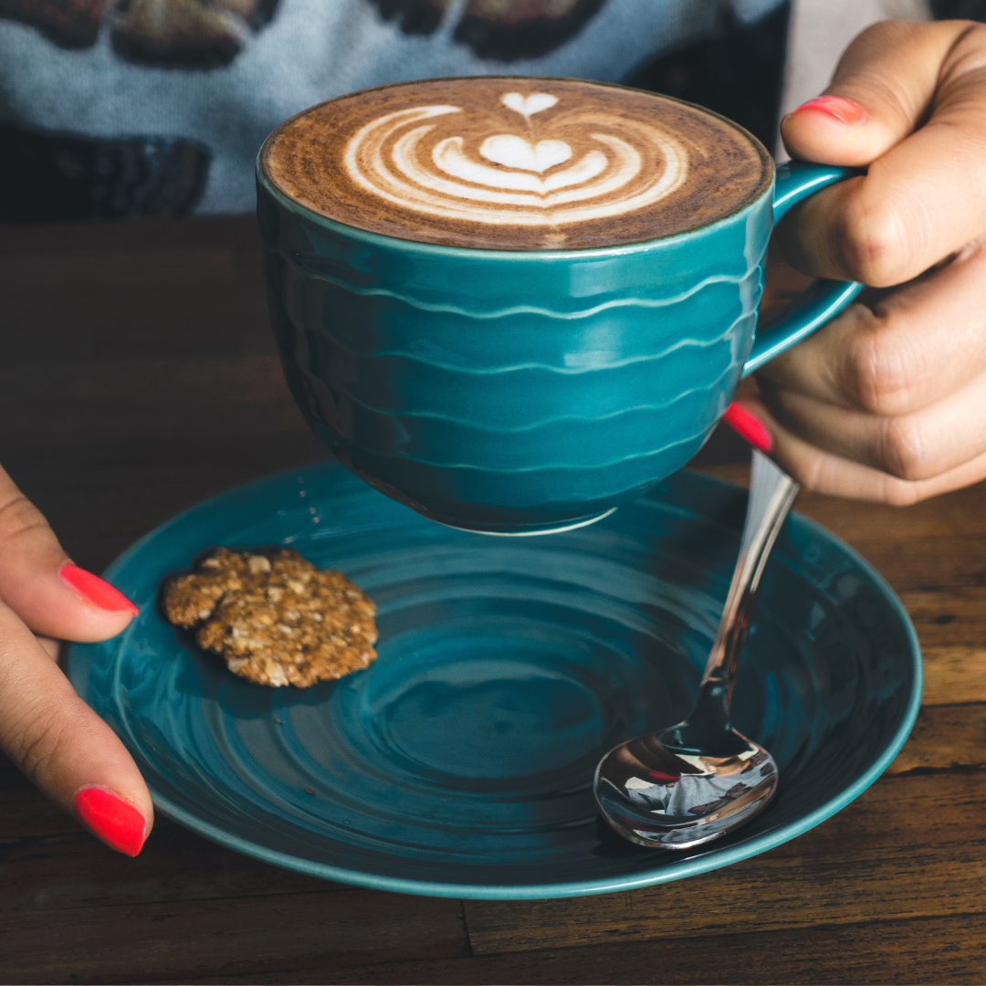 A person's hands holding a cup of Jamaican coffee in a blue cup with a cookie on the cup's saucer