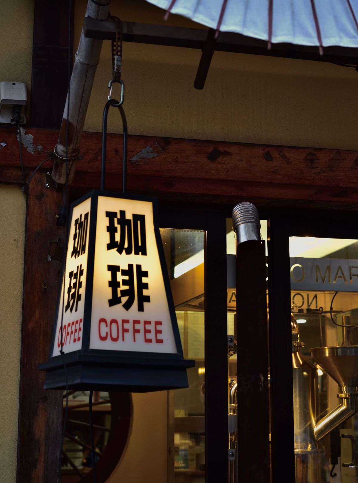 Traditional Japanese coffee shop entrance featuring a hanging lantern with Japanese characters and the word “COFFEE,” beneath a large blue paper parasol, with coffee roasting equipment visible inside.