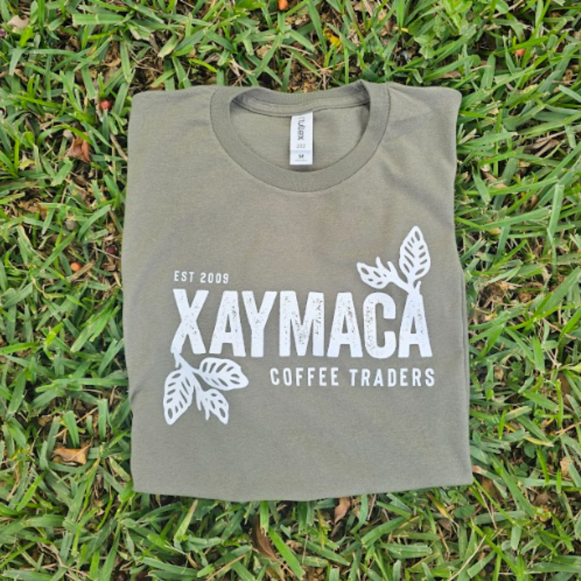 A green t-shirt with the Xaymaca Coffee Traders logo printed in the center, laid out on a grassy surface.