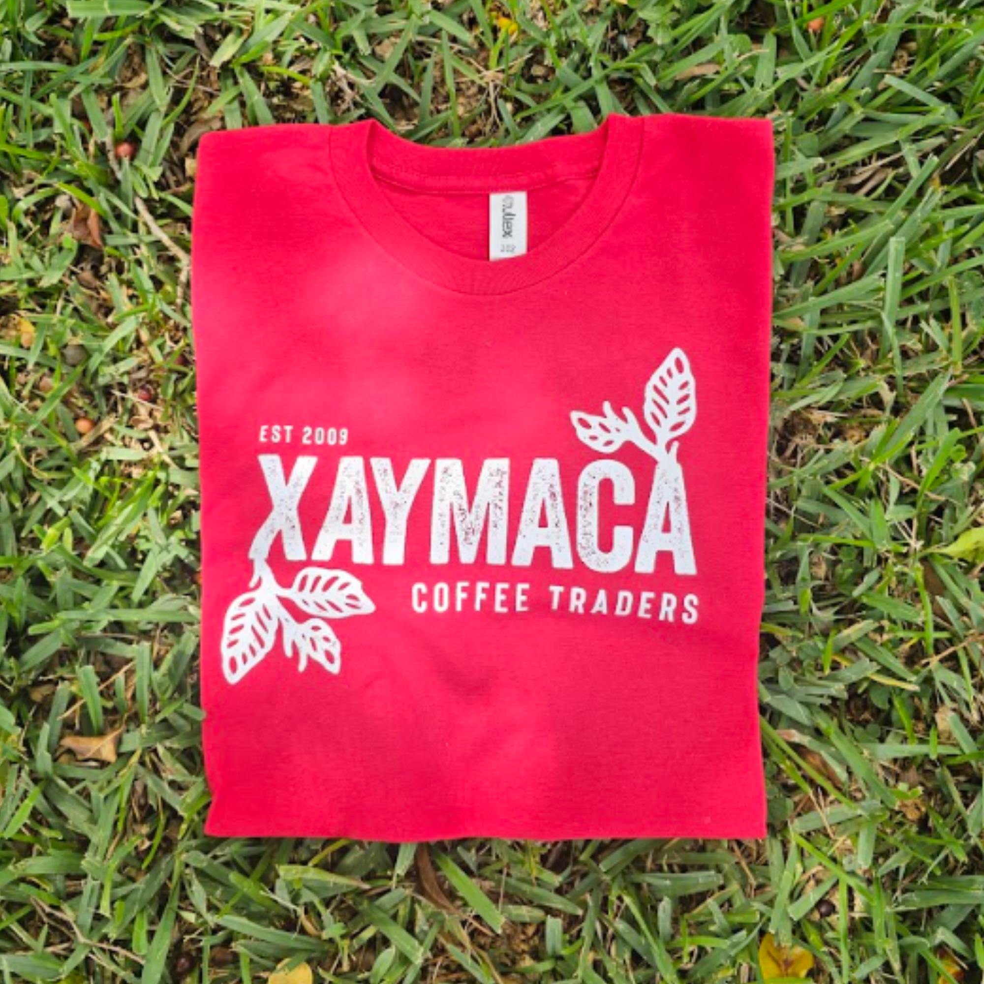 A red t-shirt with the Xaymaca Coffee Traders logo printed in the center, laid out on a grassy surface.