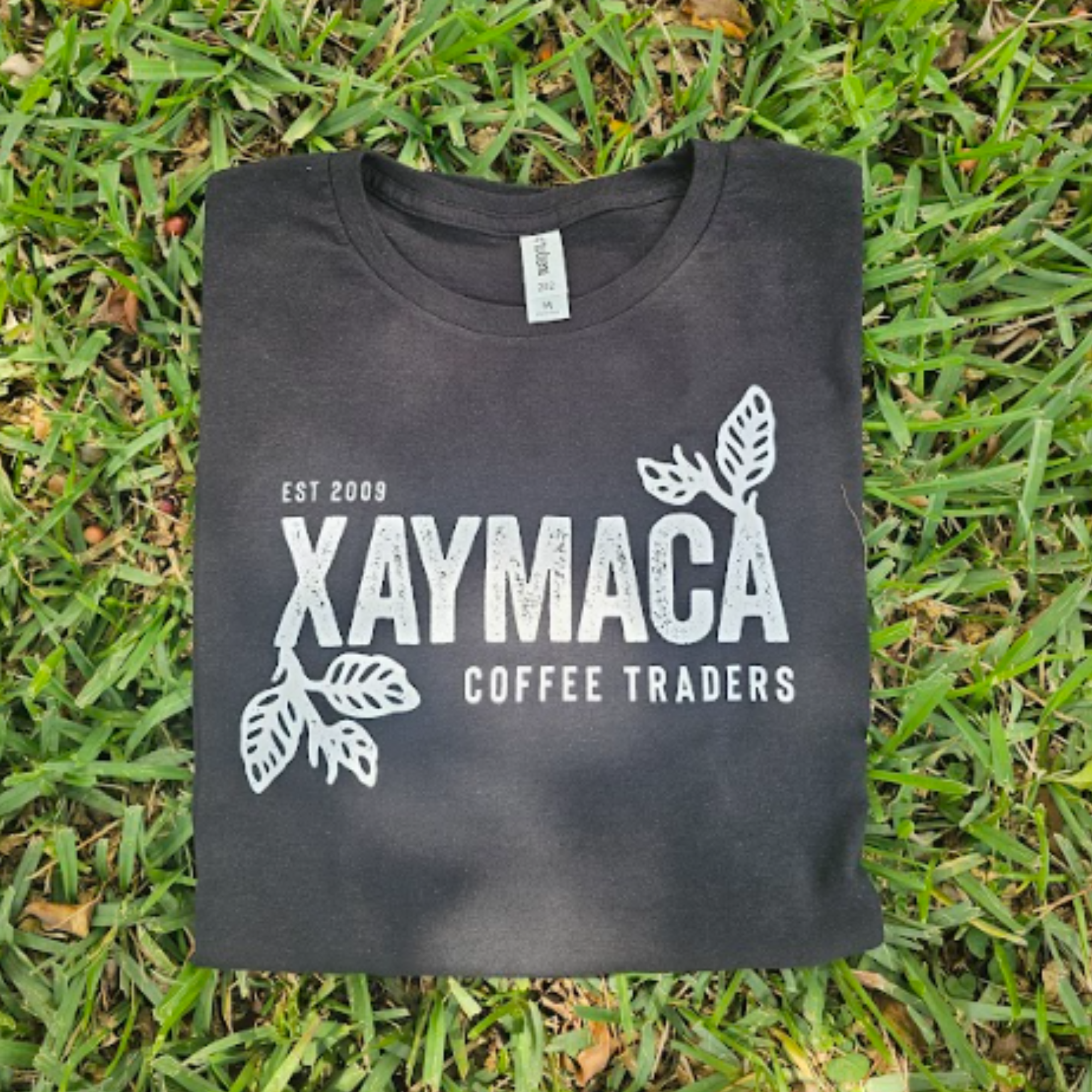 A black t-shirt with the Xaymaca Coffee Traders logo printed in the center, laid out on a grassy surface.