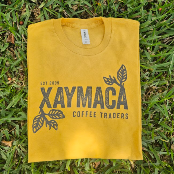 A yellow t-shirt with the Xaymaca Coffee Traders logo printed in the center, laid out on a grassy surface.