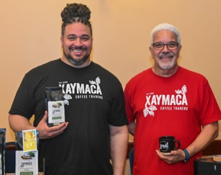 Photo of David and Damian Levy of Xaymaca Coffee Traders smiling. Damian is holding a bag of Jamaican coffee and David is holding a Xaymaca coffee mug. They are smiling.