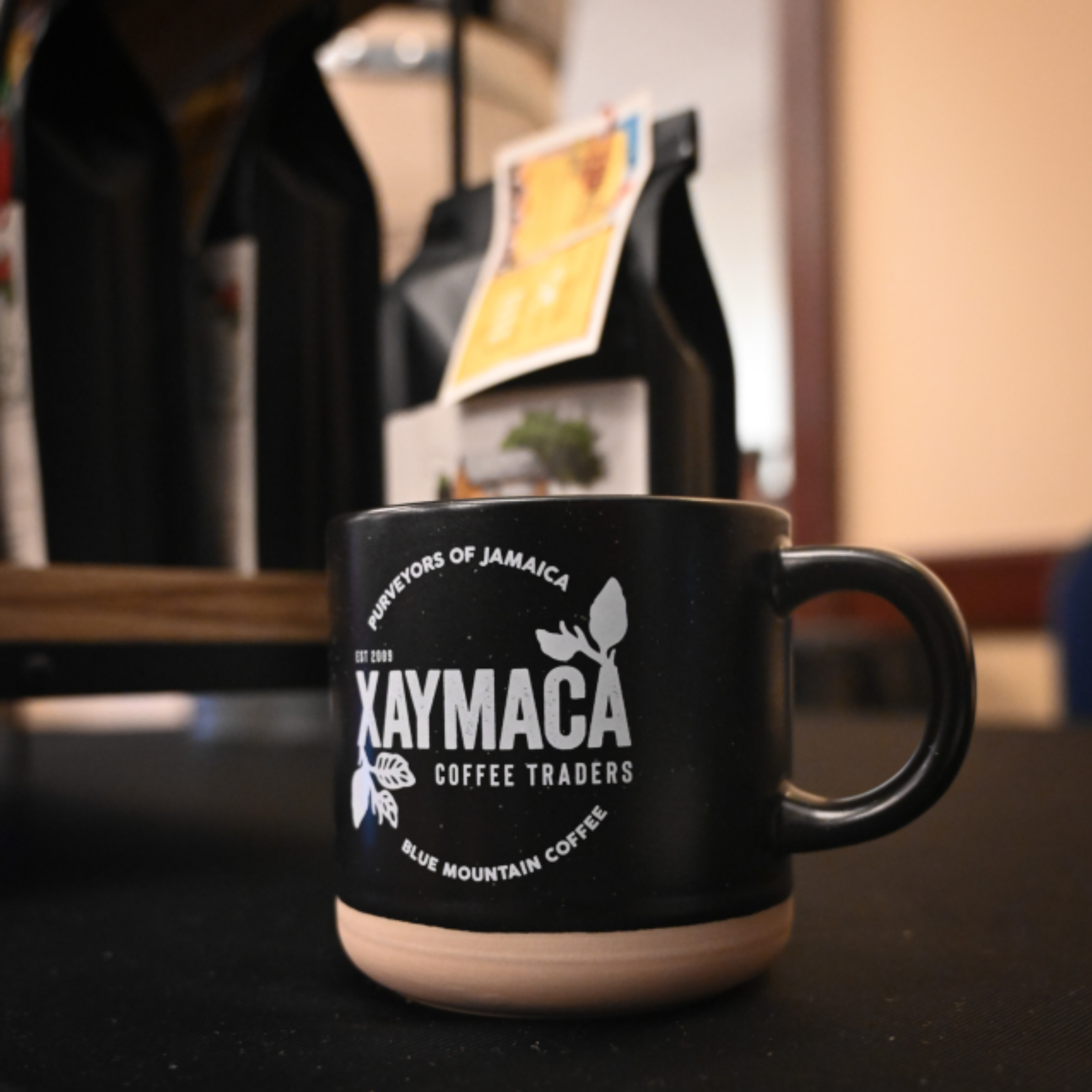 A black ceramic coffee mug with the Xaymaca Coffee Traders logo, situated on a wooden surface with coffee bags in the background.