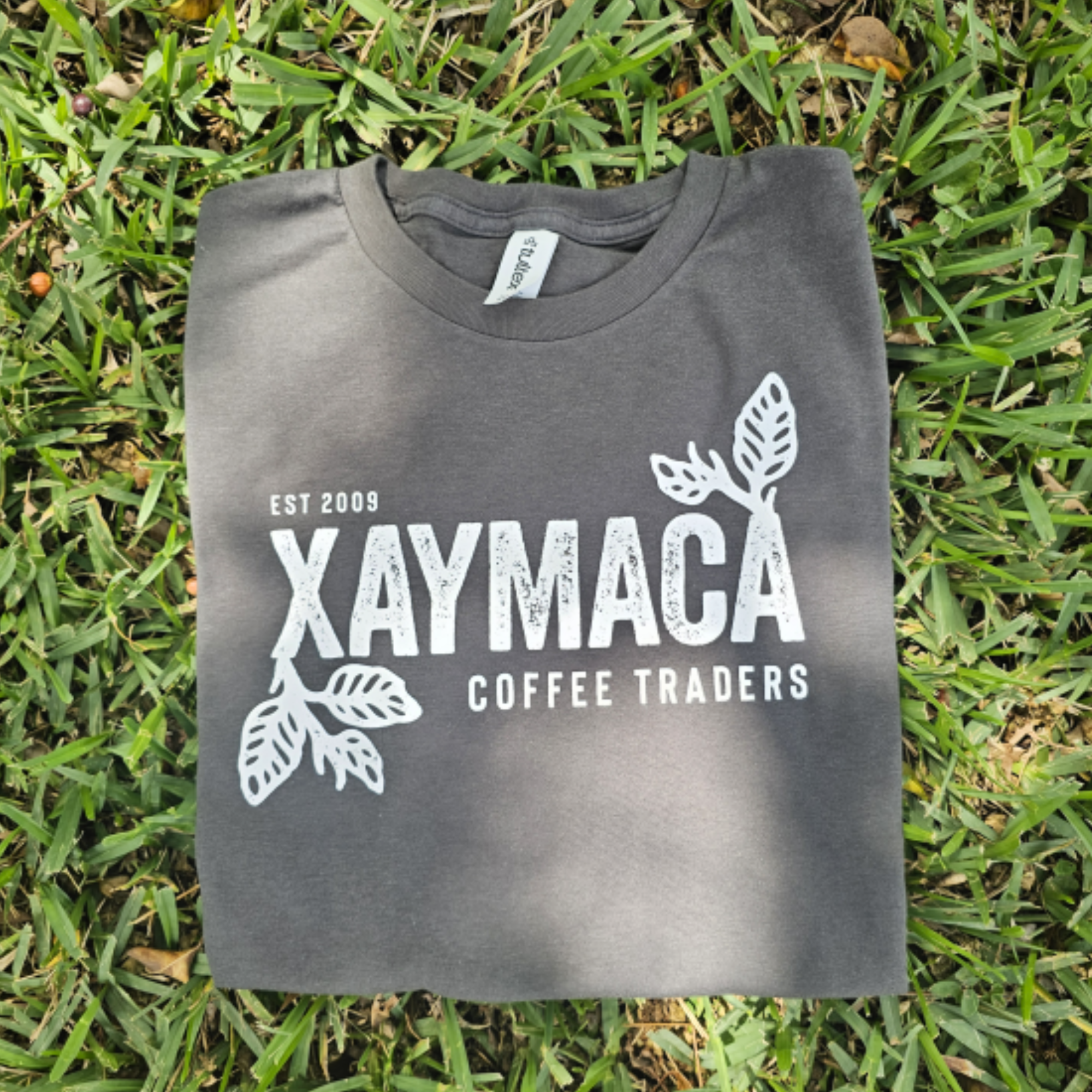 A grey t-shirt with the Xaymaca Coffee Traders logo printed in the center, laid out on a grassy surface.